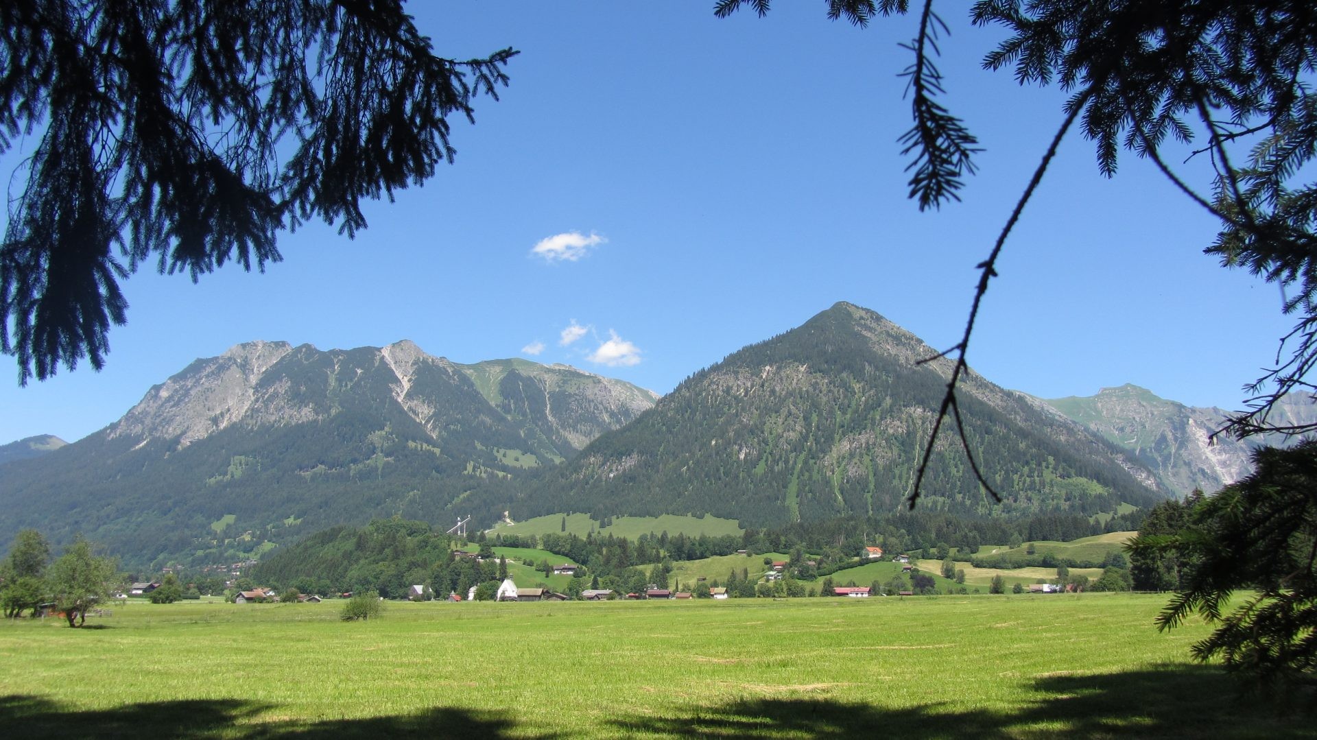 Eine Talwiese mit Höfen vor bewaldeten Bergen und einer Skisprungschanze.