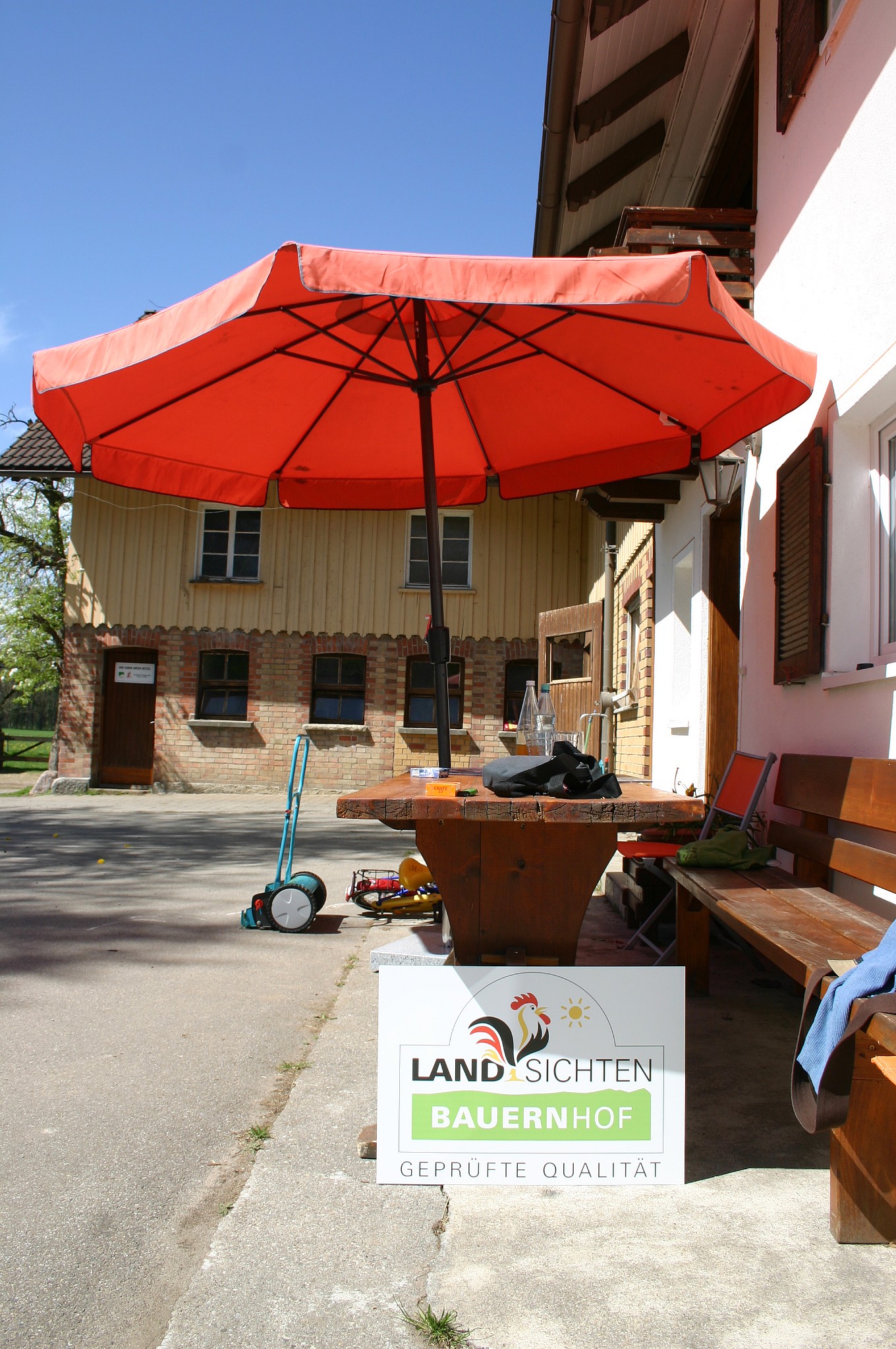 Sitzplatz mit rotem Sonnenschirm und Holzbank vor dem Bauernhaus, davor ein Schild für geprüfte Bauernhof-Qualität.