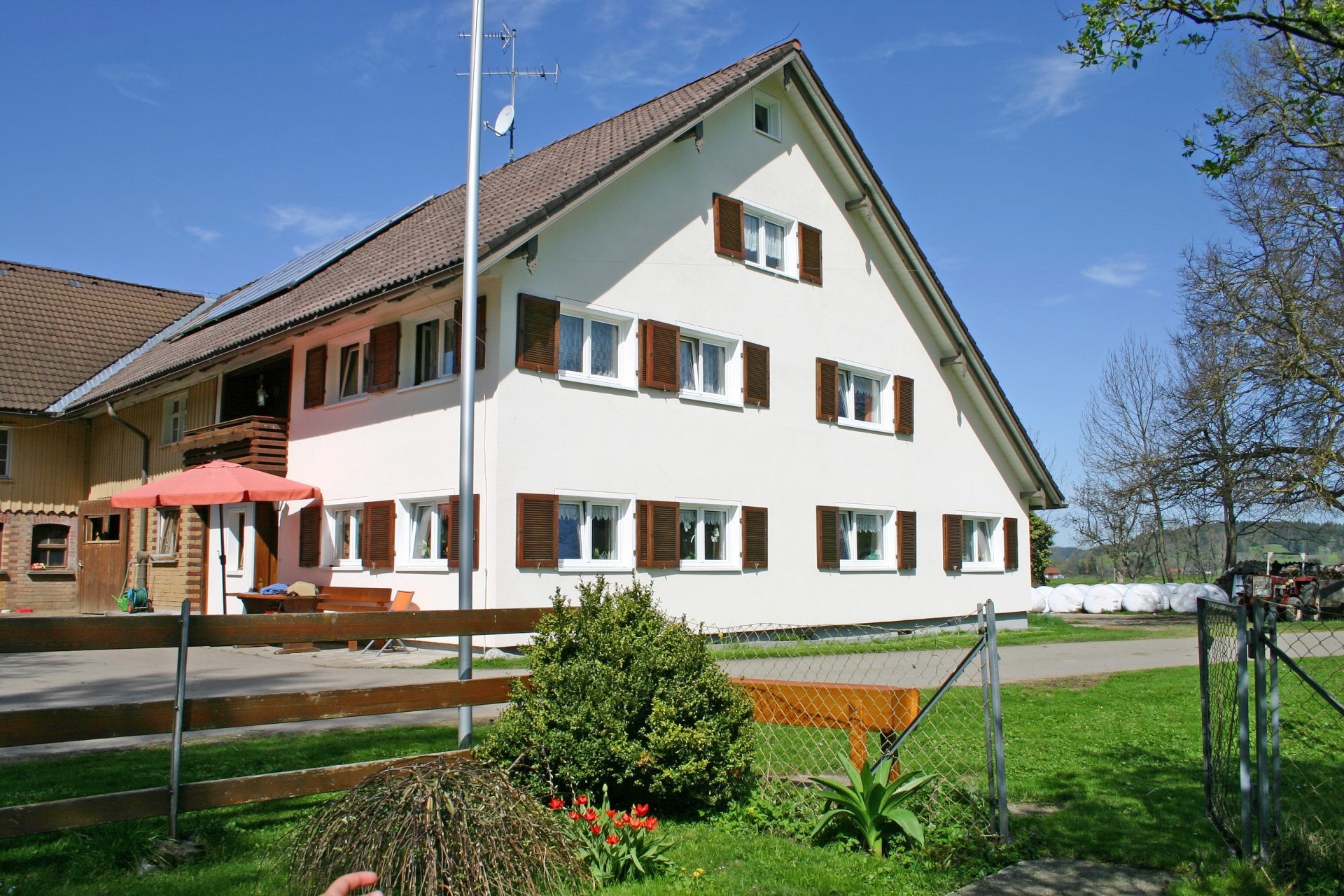 Das weiße Wohnhaus des Ferienhofs mit braunen Fensterläden, Terrasse mit Sonnenschirm und Vorgarten.