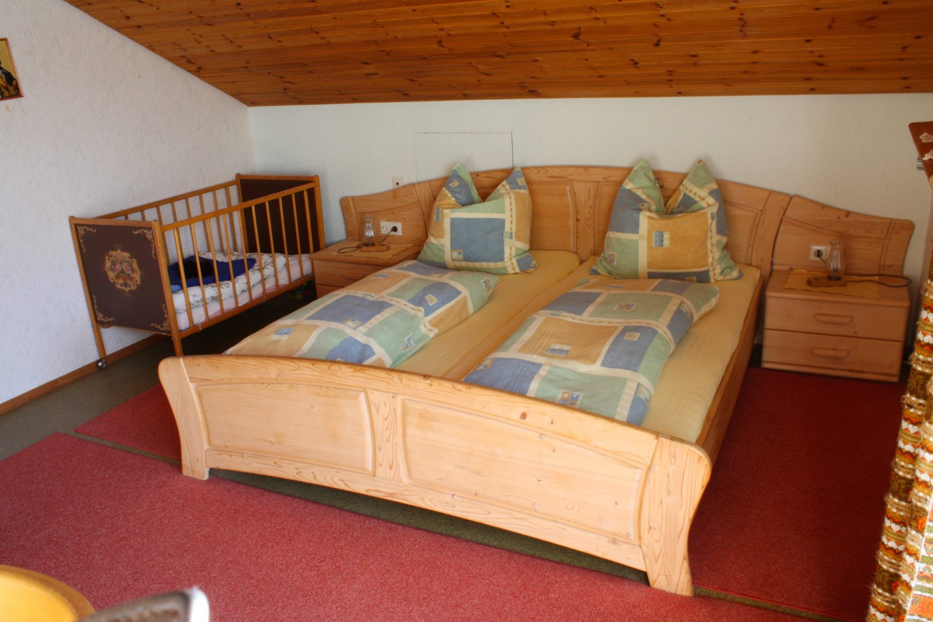 Schlafzimmer mit Doppelbett aus hellem Holz, zwei Nachttischen und einem Kinderbett unter der Dachschräge.