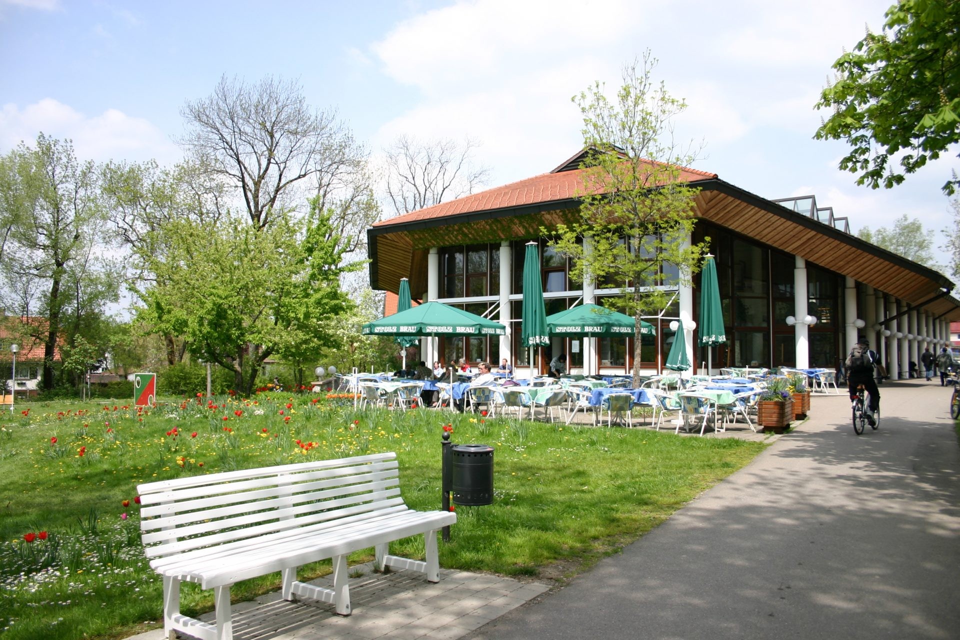 Café-Terrasse mit grünen Sonnenschirmen vor einem Gebäude im Park, davor Tulpenwiese und weiße Bank.
