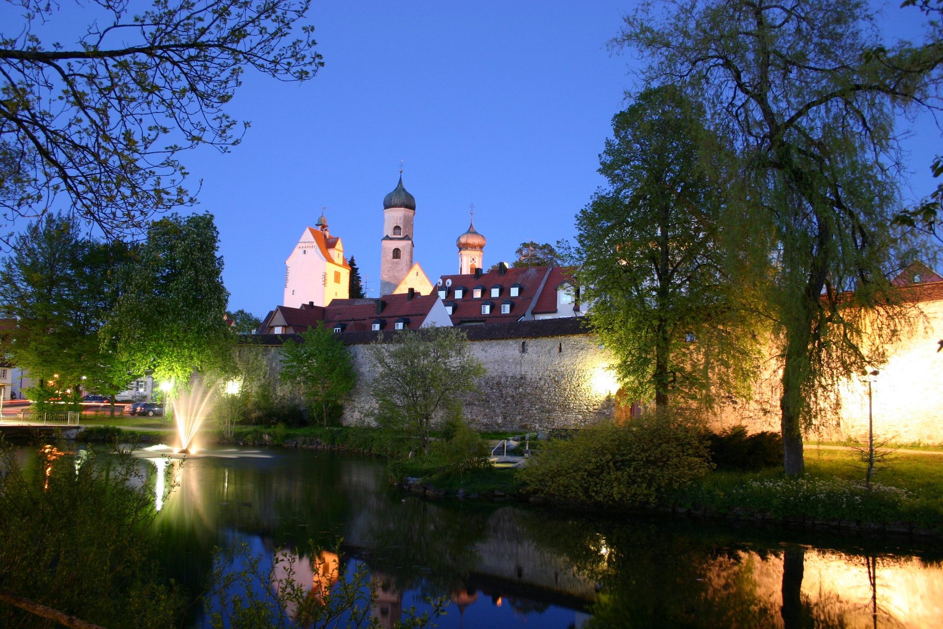 Die beleuchtete Stadtmauer von Isny mit Kirchtürmen spiegelt sich am Abend im Weiher.