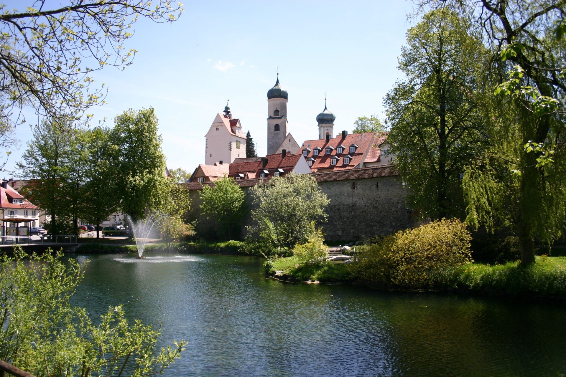 Die historische Stadtmauer von Isny mit Kirchtürmen, davor ein Weiher mit Fontäne.
