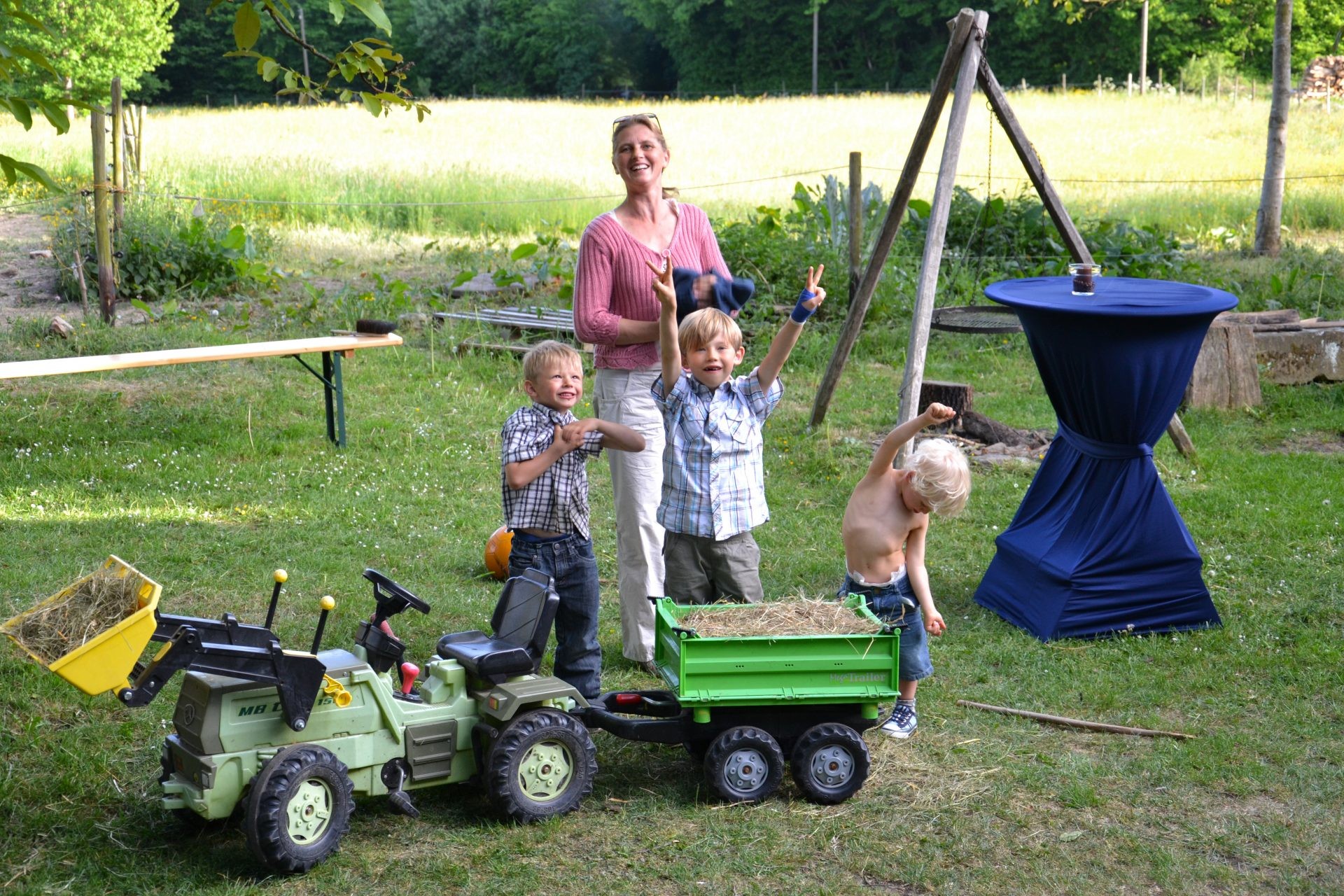 Drei Jungen und eine Frau jubeln neben einem Trettraktor mit Heuanhänger auf der Wiese.