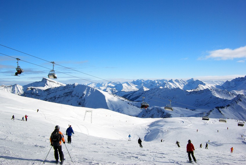 Skifahrer auf einer Piste unter einem Sessellift vor verschneiten Gipfeln.